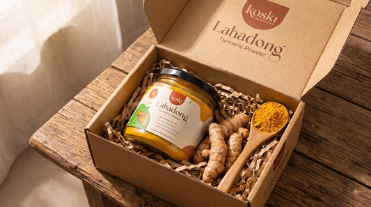 An open kraft gift box labeled Koski Organics Lakadong Turmeric Powder sits on a rustic wooden table. Inside the box is a glass jar of turmeric powder, fresh turmeric roots, and a small wooden spoon filled with bright yellow turmeric powder.