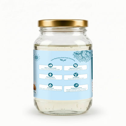 A clear glass jar of coconut oil with a gold metal lid and a light blue label showing product benefits, set against a plain white background.