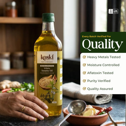 A bottle of Koski Organics White Sesame Oil stands on a kitchen counter with a hand resting near the base. The label is visible, and a promotional text panel on the right lists quality checks such as heavy metals, moisture control, aflatoxin testing, and purity verification.