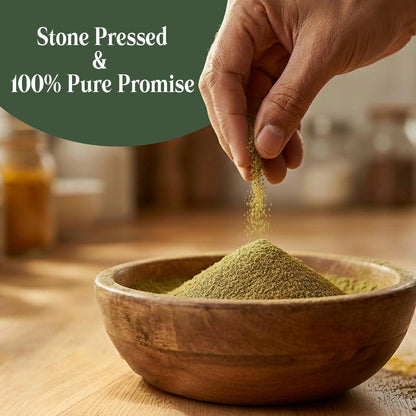 A hand sprinkles fine green coriander powder of koski organics into a wooden bowl on a wooden table, with a dark green text overlay reading 'Stone Pressed & 100% Pure Promise'.