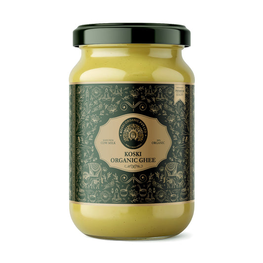A glass jar of Koski Organics Ghee with a dark green lid and a dark green label featuring intricate illustrations, containing a pale yellow ghee, set against a plain white background.
