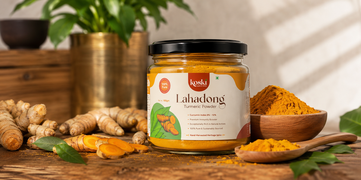 A jar of Lakadong Turmeric Powder by Koski Organics, styled on a rustic wooden table with fresh turmeric roots, a wooden spoon of powder, and a small bowl of turmeric in the background.