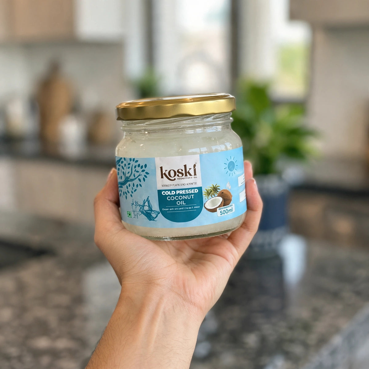 A hand holding a glass jar of kosKi Organics cold pressed coconut oil with a gold lid, set against a blurred kitchen background.
