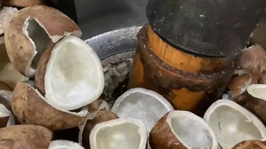 Close-up of raw coconuts, including one cracked open to show the white inner flesh, with a wooden and metal mortar-and-pestle tool in the background.