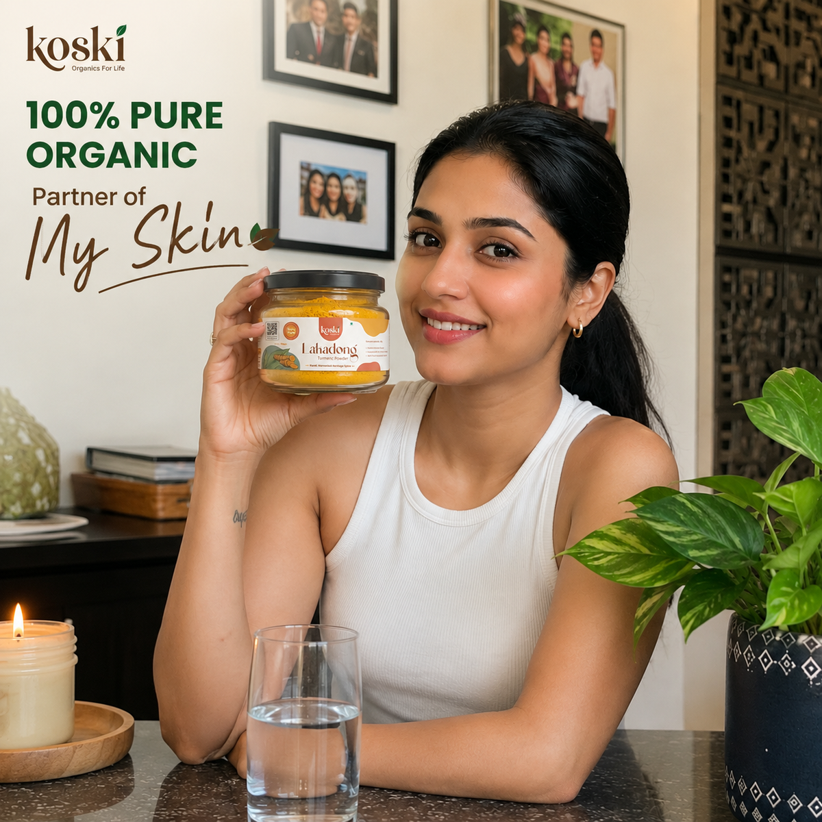 A woman in a white tank top holds up a jar of Koski Laborking Pure Organic Turmeric Paste while sitting at a table with a glass of water and a lit candle. The background shows framed family photos on a beige wall.